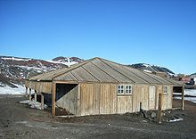 Com estrutura em madeira com porta e duas janelas pequenas. Para a esquerda é um aberto alpendre. No fundo são as montanhas parcialmente coberto de neve.