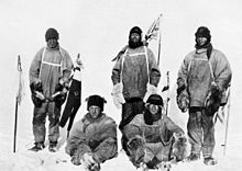 Cinco homens (três permanentes, dois sentados no chão gelado) em roupas polar pesado. Tudo parece exausta e infeliz. Os homens de pé estão carregando flagstaffs e uma bandeira da União moscas de um mastro no fundo. O partido de Scott no Pólo Sul. Da esquerda para a direita: Oates; Bowers; Scott; Wilson; Evans