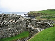 Uma pedra parede semi-circular em pistas à esquerda na existência de um edifício grande e antiga e à direita estão as ruínas de várias outras estruturas de pedra. No fundo de um penhasco baixo divide um corpo de água a partir de campos gramados.