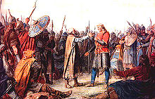 Um grupo de guerreiros em traje medieval cercar dois homens cujas posturas sugerem que eles estão prestes a abraçar. O homem à direita é mais alto, tem cabelo comprido justo e veste uma túnica vermelha brilhante. O homem à esquerda sua careca com cabelo grisalho e uma barba branca. Ele veste um longo manto marrom.
