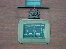 Uma tabuleta de pedra, inscrito com a imagem de um campo de futebol e vários nomes. Ele encontra-se rodeado por uma borda de pedra em forma de um estádio de futebol. Acima do tablet é uma escultura de madeira de dois homens que prendem uma grande coroa de flores.