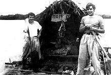 fotografia preto e branco de dois homens em uma jangada, equipado com uma grande cabana. A margem do rio é visível ao longe