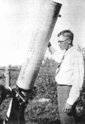 Um jovem em seus vinte e poucos anos, o uso de óculos, camisa branca, gravata e calças compridas, ergue-se em um campo aberto, ao lado de um telescópio newtoniano que descansa na terra e inclinou em direção ao céu. O telescópio é mais alto do que ele, e é cerca de oito polegadas de diâmetro. Sua mão direita está descansando em cima do barril, e ele parece um pouco passado o telescópio, para a esquerda.