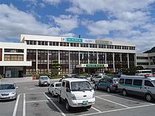 Na frente de um edifício baixo e marfim, um parque de estacionamento e os carros são mostrados.