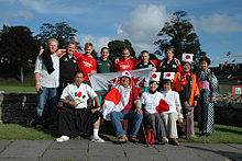 Um grupo de treze partidários levantam junto, alguns vestindo camisas de rugby enquanto outros ostentam trajes tradicionais japoneses e bandeiras japonesas.