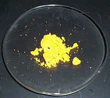 Um pouco de pó amarelo-canário senta-se, na maior parte em pedaços, num vidro de relógio de laboratório.