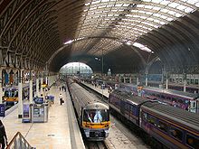 O interior de uma grande estação ferroviária com um telhado curvo suportado por vigas de ferro, apoiada por colunas de ferro, quatro trens a diesel que estão nas plataformas, os passageiros nas plataformas, na luz do dia distância pode ser visto a cena e é iluminado por luz natural através da seção central do telhado