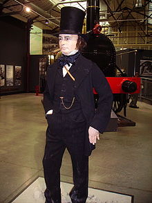 Dentro de um grande edifício, uma figura em tamanho natural de um homem em seus quarenta e tantos anos, vestido com jaqueta século XIX, calça e colete, com um proeminente corrente do relógio em seu peito. Ele usa um chapéu fogão-pipe negro alto, tem longas costeletas e tem um charuto na boca. Uma locomotiva a vapor velha no fundo.