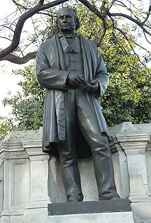 Uma escultura de bronze de metal de um homem do século XIX, vestindo um longo casaco ou casaco, calça, colete, com as ferramentas do relator nas suas mãos