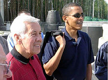 Homem de cabelos grisalhos e Obama estão, vestindo camisas pólo casuais. Obama usa óculos e tem algo pendurado no ombro direito.