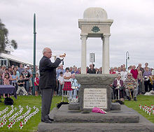Foto de um memorial ANZAC com um homem idoso que joga um cornetim. Fileiras dos povos estão sentados atrás do memorial. Muitas pequenas cruzes brancas com papoilas vermelhas ter sido preso no gramado em fileiras em cada lado do memorial.