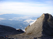 Uma vista do pico de baixa, com um pico menor à direita da foto, com montanhas arborizadas no fundo