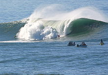 Foto de onda que quebra porte mais alto do que a humana com vários surfistas assistindo em primeiro plano