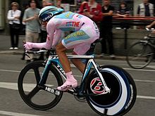 Um homem em roupas cor de rosa e sapatos cor de rosa, sentando-se abaixou-se em uma bicicleta. No fundo as pessoas estão assistindo.