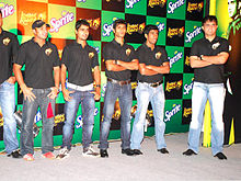 Um grupo de homens de pé, vestindo camisetas pretas e azul jeans desbotados. Todos eles estão olhando para a esquerda da imagem. The backdrop has alternate black and green boxes.
