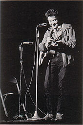 Dylan com sua guitarra no palco, rindo e olhando para baixo.