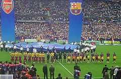 Duas equipes de desportistas, uma em camisas amarelas e um em camisas vermelhas e azuis, estão em uma linha. Na frente da linha é uma linha de crianças em camisas equivalentes, mas cada um na frente do time adversário. Um número de outras pessoas ficar em primeiro plano olhando. Atrás das equipes, um número de pessoas que detêm um grande pedaço circular de tecido com logotipos impressos em, e dois grandes banners pendem verticalmente de cima toda a cena.