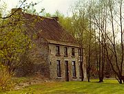 foto de uma casa de tijolos de dois andares no lado esquerdo parcialmente obscurecida por árvores com um gramado da frente e com uma fileira de árvores à direita