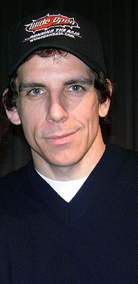 Stiller está olhando para a câmera e sorrindo. Ele está vestindo um boné de beisebol e uma camisa azul com uma t-shirt branca por baixo.