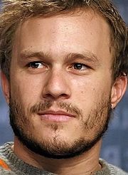 Close up do rosto de um homem com os olhos castanhos, cabelo castanho despenteado e barba desgrenhada crescimento. Ele está olhando para sua esquerda. Ele está vestindo um suéter cinza com uma faixa laranja perto de seu ombro esquerdo e braço esquerdo. O fundo é azul com fora da escrita foco.