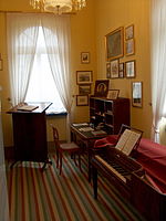 quartos decorados em estilo do início do século XIX, com o corredor listrado no assoalho; paredes pintadas de pêssego e da parede à direita pendurado com imagens pequenas; em primeiro plano o direito de um piano quadrado, em traseiro esquerdo antes de uma janela de um stand leitura, em traseiro direito uma mesa, tudo em madeira escura